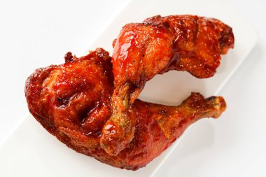 İki sulu, kahverengi ızgara tavuk bacağı, zengin bir sosla parlıyor ve beyaz dikdörtgen bir tabakta servis ediliyor. Beyaz arka planda doğal ışıkla yakalanmış..