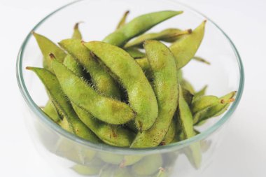 Temiz beyaz bir arka planda sergilenen canlı yeşil edamame kapsülleriyle dolu temiz bir cam kaseye yakın çekim. Popüler ve sağlıklı bir atıştırmalık..