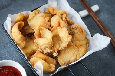 Beyaz kağıtla kaplanmış siyah bir sepetin içinde servis edilen kızarmış balık parçaları. Arka planda küçük kırmızı bir sos kasesi görünür..