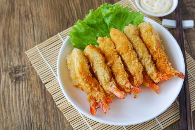 Bir tabak kızarmış karides (Ebi Fry veya Tempura), taze marulla süslenmiş ve ahşap bir plasemat ile beyaz bir tabakta servis edilmektedir. Yanında yemek çubukları ve sosu var..