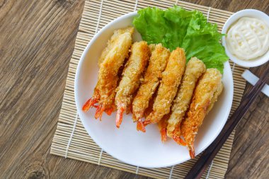 Kızarmış karides tabağının üst görüntüsü (Ebi Fry veya Tempura), taze marulla süslenmiş ve ahşap bir altlık ile beyaz bir tabakta servis edilmektedir. Yanında yemek çubukları ve sosu var..