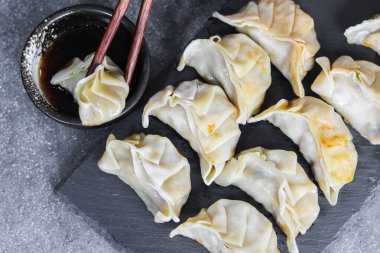 Bir tabak taze pişmiş gyoza hamur köftesi, bir tanesi yemek çubuklarıyla küçük bir tabak soya sosuna batırılmış, koyu dokulu bir yüzeyde sunuluyor..