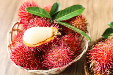 Rambutan meyveleriyle dolu canlı kırmızı dikenli deri ve yeşil yapraklarla dolu bir sepet, vücudunun yarısını gösteren bir meyve kesiği, ahşap bir arka planda sergilenir..