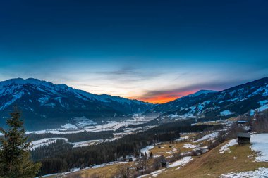 A breathtaking winter sunset near Kaprun, Austria, bathes the snow-covered mountains in golden and pink hues, as the crisp alpine air and serene lands