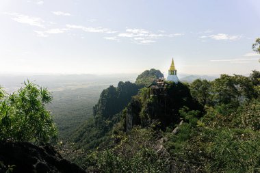 Pürüzlü, ormanlık bir dağın tepesinde, parlak, berrak gökyüzünün altında belirgin bir altın kuleye sahip beyaz bir Budist stupa (chedi) duruyor. Ziyaretçiler, stupanın bitişiğindeki ahşap izleme platformunda duruyorlar,