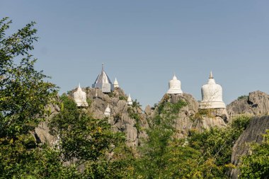 Çok sayıda beyaz ve gümüş renkli Budist pagoda (Chedis) sergilenen çarpıcı bir panorama, parlak mavi bir gökyüzünün altında birkaç sivri, kayalık kireç taşı zirvesinde tehlikeli bir şekilde duruyordu. Ön plan Lu tarafından çerçevelenmiştir.