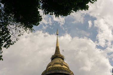 Direk olarak büyük bir Budist stupasının muhteşem altın kulesine (Chofa veya Chedi zirvesine) bakan alçak açılı manzara. Yapının yaldızlı yüzeyi parıldıyor ve sivri iğnelere dönüşüyor.