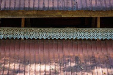 Kırsal bir binanın saçaklarına yakın plan, karmaşık bir şekilde oyulmuş ahşap kaplama ya da kaplama (delinmiş bir valans) bir bölüm üzerinde ayarlanmış, kırmızımsı-kahverengi kıvrımlı metal çatı.