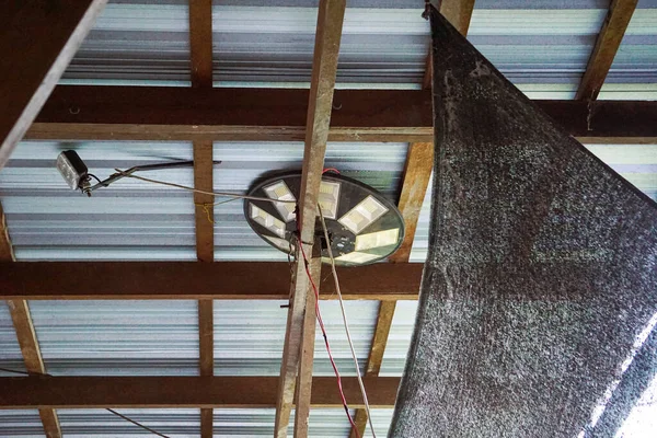 Geçici bir elektronik kurulum içeren ahşap kiriş yapısının çatı altı görüntüsü, işlevsel kırsal mühendislik ve yerli adaptasyonu vurguluyor