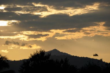Altın ışık, gün batımında siluetli bir dağ manzarası üzerinde dramatik bulutları yararak sakin ve görkemli bir doğal manzara yaratıyor..