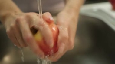 Mutfakta musluk suyunun altında kırmızı bir elmayı yıkayan ellerin yakın çekimi. Sağlık ve hijyen için ideal. Modern mutfakta kadın musluk suyunun altında elma yıkıyor..