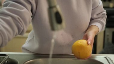  Elleri turuncu olan mutfak lavabosunda akan suyun altında. Bu, yemeden önce meyveleri yıkamanın önemini gösteriyor. Toprak ve mikropları etkin bir şekilde temizleyip daha sağlıklı hale getirmek için..
