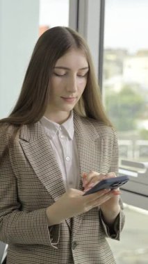Takım elbiseli kendine güvenen profesyonel bir kadın. Pencerenin yanındaki cep telefonuna mesaj yazıyor.