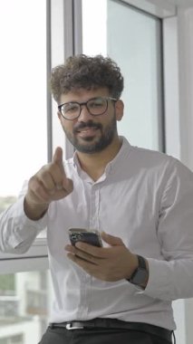 Akıllı telefonunu tutarken kameraya gülümseyen ve işaret eden olumlu genç bir erkek.