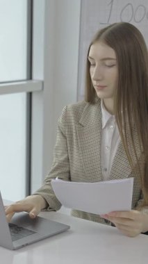 Ofiste çalışan, dizüstü bilgisayarda yazan ve önemli evrakları kontrol eden genç bir kadın.