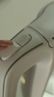 Mavi ışıklı beyaz bir su ısıtıcısının güç düğmesine basan bir kadının yakın çekimi.