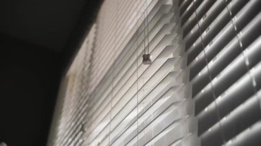 Pencerede beyaz yatay bir külüstür açılır ve güneş ışığı karanlık bir odaya girer.