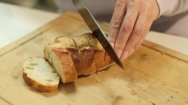 Kadınların elleri taze bir somun ekmeği kesiyor. Mutfakta tahta bir kesme tahtasının üzerinde bıçakla.