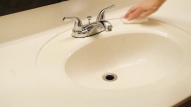 Beyaz seramik bir banyo lavabosu ve krom musluğu beyaz bir süngerle silen insan eli.