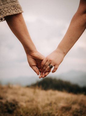 Birbirlerini bir dağ manzarasına karşı tutan iki elin yakınlaşması, aşkı, şefkati ve duygusal bağı simgeliyor. Sahne, açık hava ortamında romantizm, güven ve beraberliği yansıtıyor..