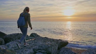 Genç bir kadın denize doğru yürüyor, saçlarını yavaş çekimde düzeltiyor ve ufka bakıyor.