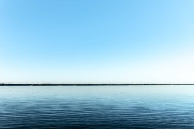 Açık mavi gökyüzünün altında sakin bir göl manzarası. Huzurlu su yüzeyi ve temiz ufuk huzurlu ve sakin bir atmosfer yaratır..