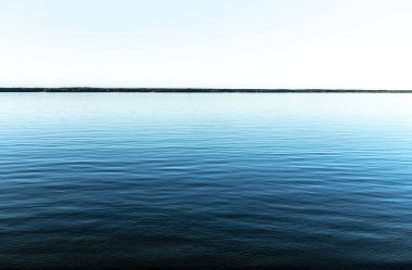 Açık mavi gökyüzünün altında sakin bir göl manzarası. Huzurlu su yüzeyi ve temiz ufuk huzurlu ve sakin bir atmosfer yaratır..