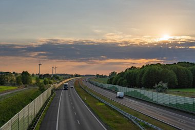 Sıcak günbatımı ışıklarıyla yıkanan bir yaz otobanı, yan tarafında bir gürültü bariyeri ve uzaktan görülebilen rüzgar türbinleri ile. Doğanın dokunuşuyla modern bir manzara..
