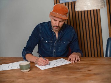 Turuncu bereli bir adam, kahve fincanıyla ahşap masadaki resimli kareler çiziyor.