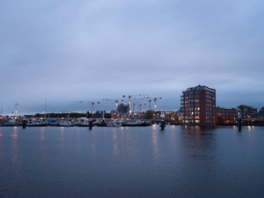 Alacakaranlıktaki yat limanında aydınlık binalar, tekneler ve aktif inşaat vinçleri suya yansıyor.