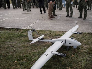 Beyaz keşif aracı otların üzerinde sergilendi ve askeri personel oluşumu asfalt zeminde görülebilir.