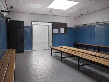 Spor salonu soyunma odası içi metal dolaplar ahşap banklar ve açık kapılı mavi fayanslı duvarlar.