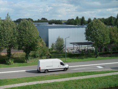 Beyaz kargo kamyonu, güneş paneli tepe örtüsü ve manzaralı modern endüstriyel tesisin yanından geçiyor.