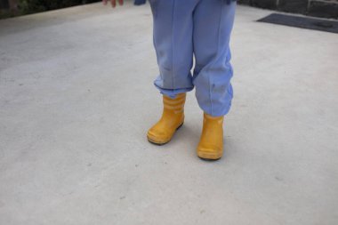 Mavi pantolonlu çocuk tarafından giyilen parlak sarı lastik çizmeler beton kaldırımda duruyor.