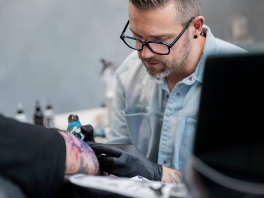 Gözlüklü profesyonel erkek dövme sanatçısı. Stüdyoda detaylı müşteri çalışmaları üzerinde yoğunlaşıyor.