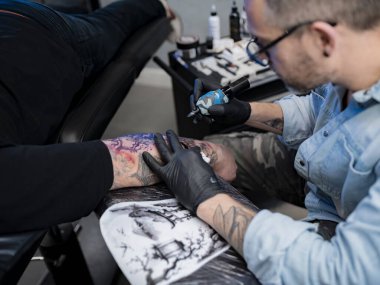 Dövmeci, müşteri kolu dövmesi üzerinde stüdyoda yan açıyla çalışıyor.