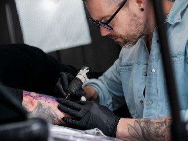 Gözlüklü erkek sanatçı renkli kol dövmesi işine odaklanmış.