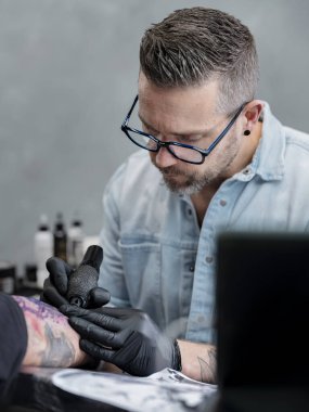 Mavi gözlüklü dövme sanatçısı, profesyonel stüdyoda müşterinin kolunda mor şablon çizgili ayrıntılı Japon kolu dövmesi üzerinde yoğunlaşıyor..