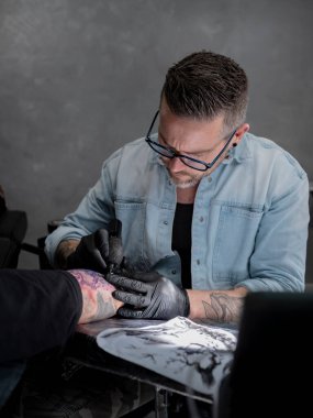 Geleneksel Japon pelerin kapağı tasarımı üzerinde çalışan dövme sanatçısı müşterinin ön kolunda görünen mor şablon hatlarıyla çalışıyor..