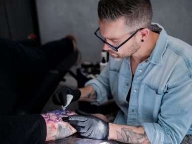 Stüdyoda müşterinin ön kolunda görünür mor şablon transferi ile Japon peyzaj kolunda çalışan dövme sanatçısının yan görüntüsü.