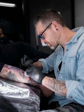 Dövme sanatçısının yan profili detaylı Japon manzara kapağı çalışmalarına odaklanıyor. Stüdyo ışıkları dramatik bir etki yaratıyor..