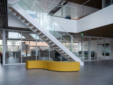 İçinde kıvrımlı sarı bank ve mimari cam merdiven bulunan modern şirket iç mimarisi.