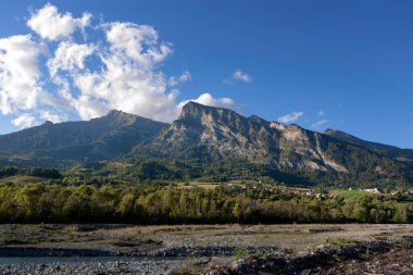 Orman yamaçları olan küçük bir köyün üzerinde dramatik dağ zirveleri yükseliyor. Kırsal yerleşim ve doğal güzelliği gösteren Avrupa dağlık manzarası.