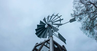 Metal yel değirmeni heykeli, ağaç dallarıyla gri gökyüzüne karşı. Klasik dekoratif rüzgar enerjisi yapısı. Anamorfik mercekle çekilmiş..