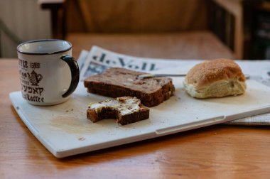 Kahve fincanı, tereyağlı ekmek dilimleri, susamlı rulo ve tahta tahtada gazete ile kırsal kahvaltı sahnesi. Yaşam tarzı ve konukseverlik içeriği için sabah rutini ve konforlu yiyecekler.