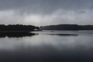 Alacakaranlıkta sakin bir göl. Küçük bir ada silueti ve orman yansıması atmosferik minimalist manzara yaratıyor.