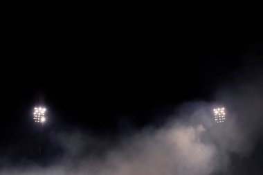 Geceleyin yoğun siste parlayan iki stadyum projektörü, duman ve sisli simetrik bileşim.