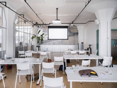 Projektör ekranlı modern toplantı odası, beyaz masalar ve sandalyeler, endüstriyel tavan ve bitkiler, eğitim alanı