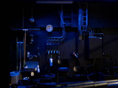 Sahne arkasındaki kulis, duvar saati, lavabo, istiflenmiş sandalyeler ve mavi ışıklandırmalı elektrik panelleri.