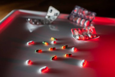 İlaç hapları ve kapsüller metal tepside etrafa saçılmış parlak kırmızı ışık ve boş su toplayıcıları.